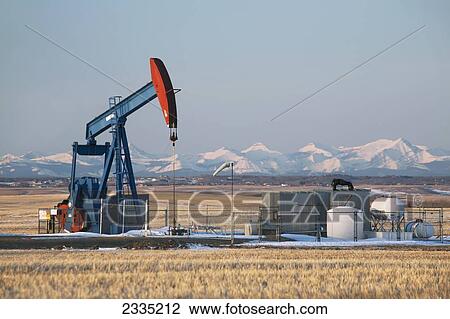 Stock Image - Pumpjack in a field with snow covered mountains and blue sky at sunrise; Alberta, Canada. Fotosearch