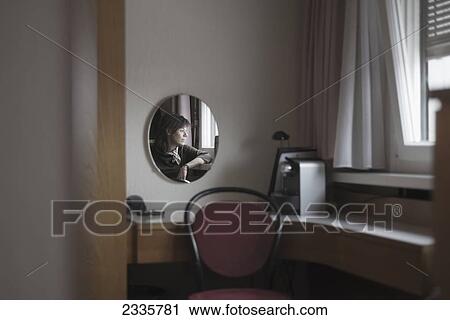 Stock Image - Reflection of a woman in a mirror as she looks out the window. Fotosearch