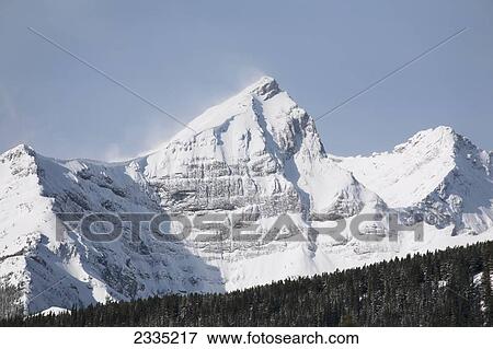 Stock Photo - Snow covered mountain range with blue sky and snow blowing off peak; Kananaskis Country, Alberta, Canada. Fotosearch