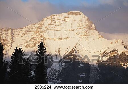 Picture - Snow peaked mountain with orange glow of sunrise with blue sky and hazy clouds; Kananaskis Country, Alberta, Canada. Fotosearch