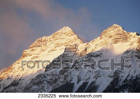 Snow peaked mountain with orange glow of sunrise with blue sky and hazy clouds; Kananaskis Country, Alberta, Canada View Large Photo Image Stock Photography - Snow peaked mountain with orange glow of sunrise with blue sky and hazy clouds; Kananaskis Country, Alberta, Canada. Fotosearch