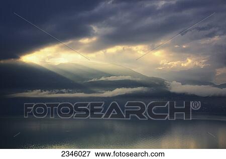 Stock Photo - The sun shining through storm clouds over water; Isole di Brissago, Ticino, Switzerland. Fotosearch