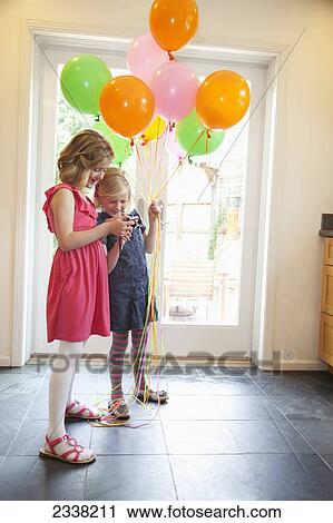 Stock Image - Two young girls standing inside a house with balloons while looking at pictures on a camera; Pacifica, California, United States of America. Fotosearch