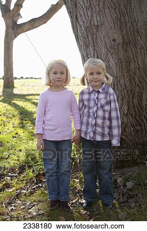 Stock Image - Two young girls with blond hair hold hands standing in front of a large tree trunk; Crab Cove, California, United States of America. Fotosearch