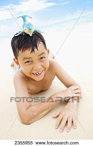 Stock Photography - A Boy Laying On The Sand At The Water's Edge With A Toy Whale On His Head; Kailua, Oahu, Hawaii, United States Of America. Fotosearch