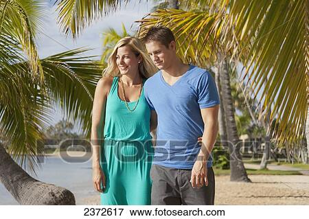 Stock Photo - A couple walk on a beach with palm trees along the water's edge; Honolulu, Hawaii, United States of America. Fotosearch