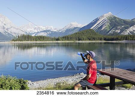 Stock Photograph - A Man Drinks From A Water Bottle On The Edge Of A Lake Overlooking The Rocky Mountains; Kananaskis, Alberta, Canada. Fotosearch
