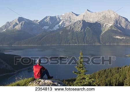 Stock Image - A Man Sits On A Rock Overlooking A Lake And The Rocky Mountains; Kananaskis, Alberta, Canada. Fotosearch