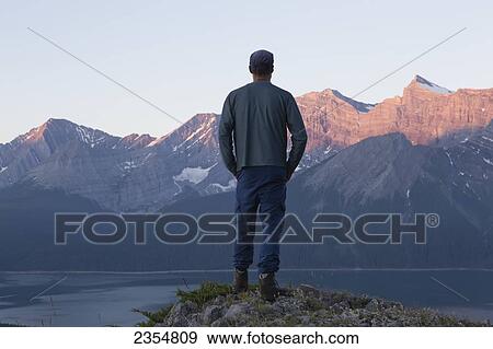 A Man Stands On A Ridge Overlooking A Lake Towards The Rocky Mountain Peaks; Kananaskis, Alberta, Canada View Large Photo Image Stock Photo - A Man Stands On A Ridge Overlooking A Lake Towards The Rocky Mountain Peaks; Kananaskis, Alberta, Canada. Fotosearch