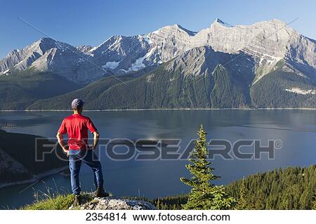 A Man Stands On A Rock Overlooking A Lake And The Rocky Mountains; Kananaskis, Alberta, Canada View Large Photo Image Picture - A Man Stands On A Rock Overlooking A Lake And The Rocky Mountains; Kananaskis, Alberta, Canada. Fotosearch