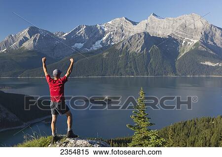 A Man Stands With Arms Raised On A Rock Overlooking A Lake And The Rocky Mountains; Kananaskis, Alberta, Canada View Large Photo Image Stock Photography - A Man Stands With Arms Raised On A Rock Overlooking A Lake And The Rocky Mountains; Kananaskis, Alberta, Canada. Fotosearch