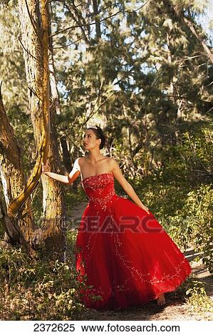 Stock Photography - A woman in a formal red gown posing by a tree; Honolulu, Hawaii, United States of America. Fotosearch