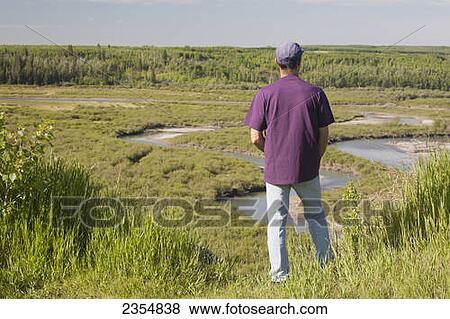 Stock Photo - Man Standing On A Cliff Overlooking A Lush River Valley With Blue Sky; Calgary, Alberta, Canada. Fotosearch