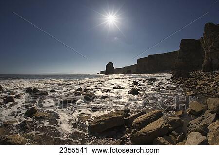 Stock Image - Water Washing Over The Rocks On Shore And Cliffs Silhouetted By A Bright Sun On Marsden Bay; South Shields, Tyne And Wear, England. Fotosearch