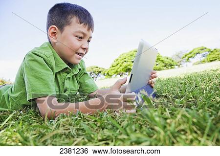 Stock Photograph - A boy laying on the grass at a park using an electronic tablet; Honolulu, Oahu, Hawaii, United States of America. Fotosearch