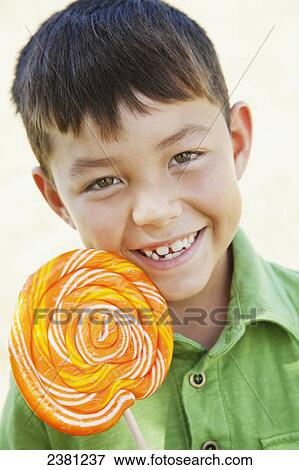 Stock Photo - A boy smiling holding a large orange lollipop candy; Honolulu, Oahu, Hawaii, United States of America. Fotosearch