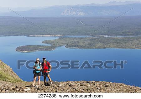 Stock Photo - A couple stands posing for a portrait with the landscape of mountains and lakes in the valley in the background; Yukon, Canada. Fotosearch