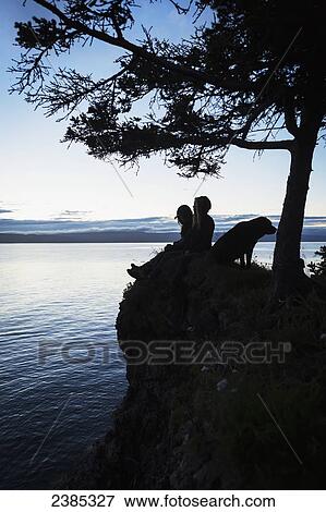 Stock Photo - A couple with a dog sit on a ridge at the water's edge looking out over Halibut Cove, Kachemak Bay, Kenai Peninsula; Alaska, United States of America. Fotosearch
