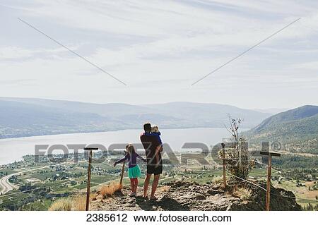 A father with his daughters standing on a rock ledge overlooking Lake Okanagan; Peachland, British Columbia, Canada View Large Photo Image Stock Image - A father with his daughters standing on a rock ledge overlooking Lake Okanagan; Peachland, British Columbia, Canada. Fotosearch