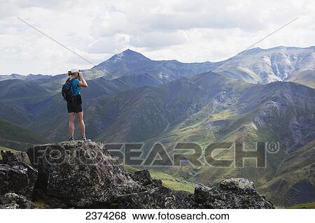 Stock Photo - A female hiker stands looking over the landscape of mountains and valleys with a camera; Yukon, Canada. Fotosearch