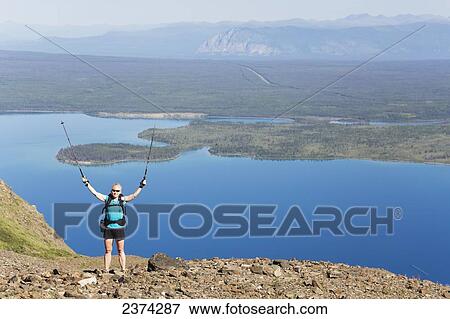 Stock Photo - A female hiker stands with arms raised looking over the landscape of mountains and lakes in the valley; Yukon, Canada. Fotosearch
