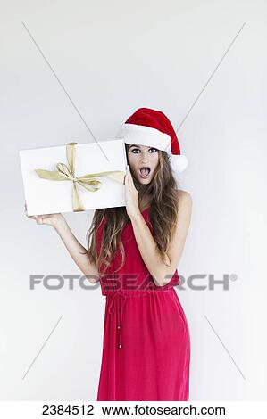 Stock Image - A girl dressed in a red shirt and santa hat holds a wrapped gift with a surprised look on her face; Hawaii, United States of America. Fotosearch