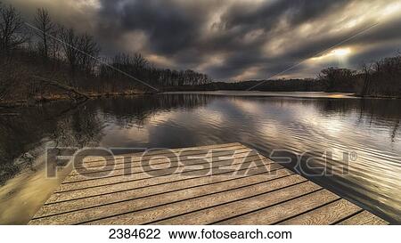 Stock Image - A God ray bursts open with a dock and stunning reflections on the water; Whitnall Park, Wisconsin, United States of America. Fotosearch