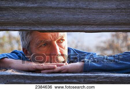 Stock Photograph - A man views the sunset through fence boards on Mount Tzouhalem on Vancouver Island; British Columbia, Canada. Fotosearch