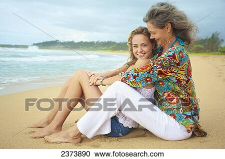 Stock Image - A mother and daughter sitting together on the beach at the water's edge; Kauai, Hawaii, United States of America. Fotosearch