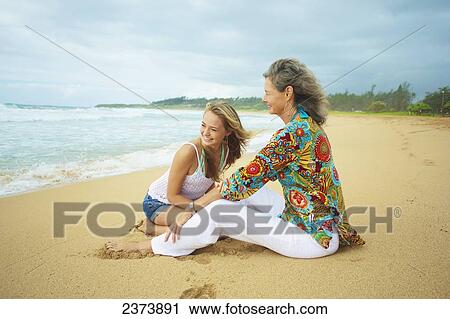 Stock Image - A mother and daughter sitting together on the beach at the water's edge; Kauai, Hawaii, United States of America. Fotosearch