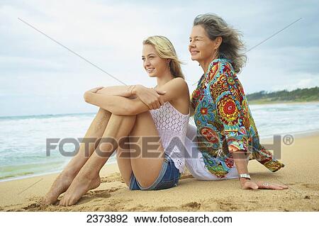Stock Image - A mother and daughter sitting together on the beach at the water's edge; Kauai, Hawaii, United States of America. Fotosearch