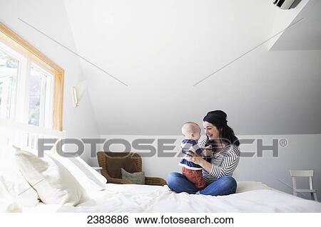Stock Photograph - A mother plays with her infant son on a bed; Victoria, British Columbia, Canada. Fotosearch