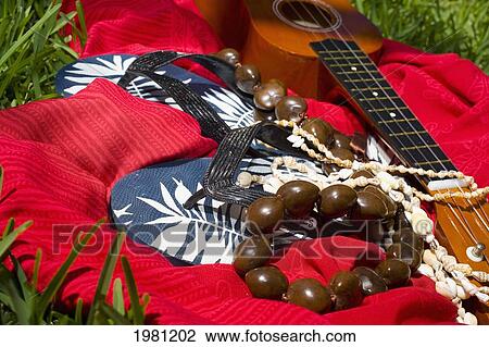 Stock Image - A Pair Of Flipflops With Kukui Nut And Shell Leis And Ukulele Laying On A Red Cloth In The Grass.. Fotosearch