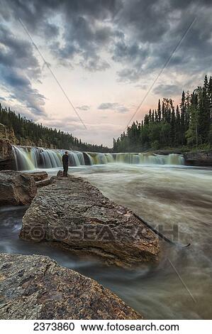 Stock Image - A person watches as the Trout River flows over Sambaa Deh Falls in Sambaa Deh Territorial Park; Northwest Territories, Canada. Fotosearch