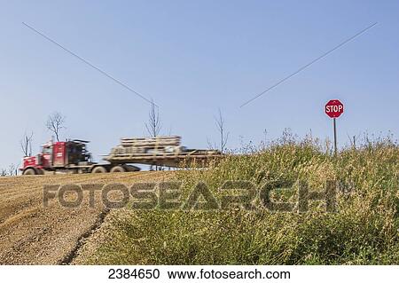 Stock Image - A semi truck travels on the highway past the stop sign and intersection to a gravel road; Fort McKay, Alberta, Canada. Fotosearch