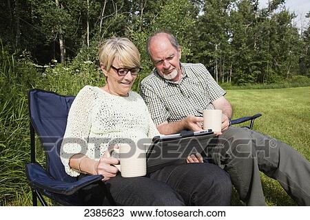 Stock Image - A senior couple sitting together in lawn chairs at a park looking at a tablet; Stony Plain, Alberta, Canada. Fotosearch