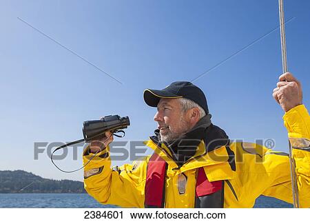 Stock Image - A senior man with binoculars sailing near Vancouver Island; British Columbia, Canada. Fotosearch
