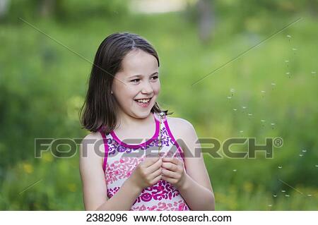 Stock Photograph - A smiling young girl holding dandelion seeds blowing in the wind in the summertime; Palmer, Alaska, United States of America. Fotosearch