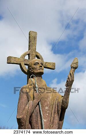 A statue of an historic figure standing against a celtic cross holding a staff; Holy Island of Lindisfarne, Northumberland, England View Large Photo Image Picture - A statue of an historic figure standing against a celtic cross holding a staff; Holy Island of Lindisfarne, Northumberland, England. Fotosearch