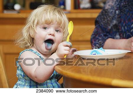 Stock Image - A toddler eats cake with blue icing and shows her blue tongue; Alberta, Canada. Fotosearch