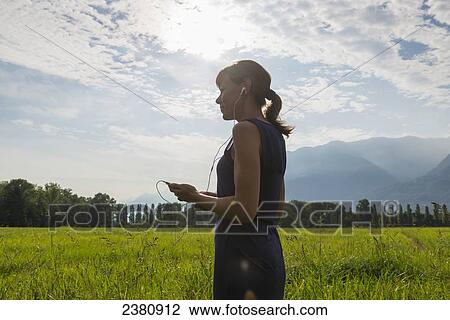 Stock Image - A woman standing in a field in the sunlight listening to music through earbuds; Locarno, Ticino, Switzerland. Fotosearch