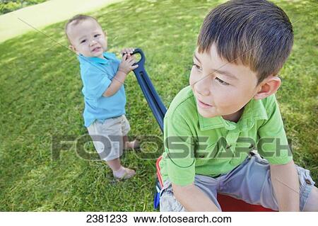 Stock Image - A young boy pulling a wagon in the park with his big brother in it; Honolulu, Oahu, Hawaii, United States of America. Fotosearch