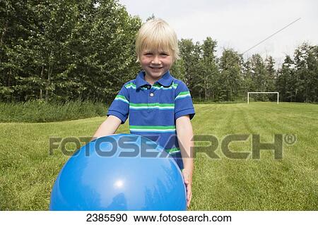 Stock Image - A young boy with blond hair plays with a large blue ball on the grass; Stony Plain, Alberta, Canada. Fotosearch