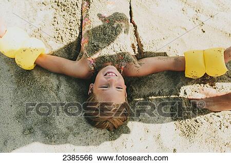 Stock Photograph - A young girl being buried in the sand on the beach; Peachland, British Columbia, Canada. Fotosearch