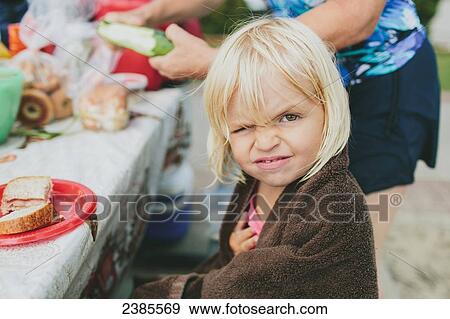 Stock Photo - A young girl winking at the camera as she sits at a picnic table for a meal; Peachland, British Columbia, Canada. Fotosearch