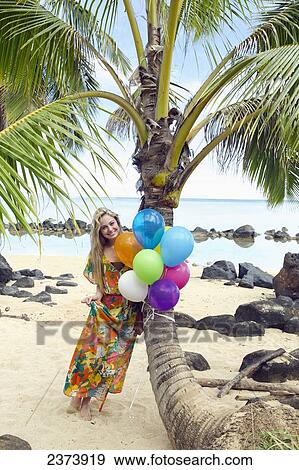 Stock Photo - A young woman in a floral dress holding a bundle of colourful balloons on the beach; Kauai, Hawaii, United States of America. Fotosearch