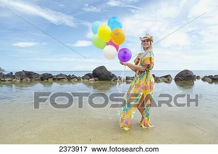 A young woman in a floral dress standing on the beach holding a bundle of colourful balloons; Kauai, Hawaii, United States of America View Large Photo Image Stock Photo - A young woman in a floral dress standing on the beach holding a bundle of colourful balloons; Kauai, Hawaii, United States of America. Fotosearch