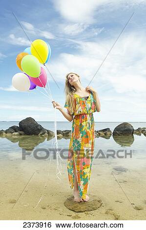 Stock Photograph - A young woman in a floral dress standing on the beach holding a bundle of colourful balloons; Kauai, Hawaii, United States of America. Fotosearch