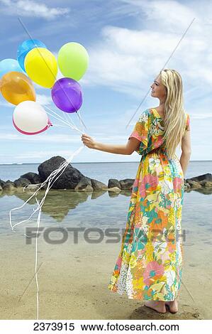 A young woman in a floral dress standing on the beach holding a bundle of colourful balloons; Kauai, Hawaii, United States of America View Large Photo Image Stock Photography - A young woman in a floral dress standing on the beach holding a bundle of colourful balloons; Kauai, Hawaii, United States of America. Fotosearch