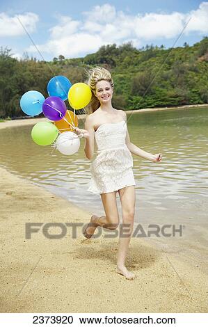 Stock Image - A young woman runs on the beach with a bundle of colourful balloons; Kauai, Hawaii, United States of America. Fotosearch